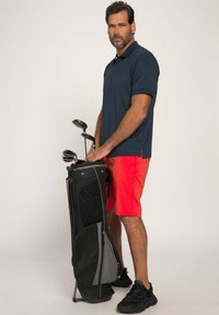 Golfbag met zwart en grijze accenten, vasthoudend een set clubs. Model draagt een donkerblauwe polo en felrode shorts, staande in zwarte sportschoenen.