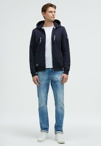 Sudadera azul marino con detalles en blanco y cordones, con cremallera, bolsillos delanteros y un parche de logo. La llevan con jeans de mezclilla de color azul claro y zapatillas blancas.