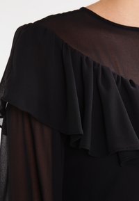 Blouse en tissu noir transparent avec un grand détail volanté sur l'épaule et le haut de la poitrine, portée par une personne.