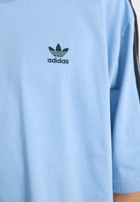 Šviesiai mėlyna medvilninė marškinėliai su trumpomis rankovėmis, su juodu Adidas logotipu ant krūtinės ir juodais akcentais šonuose.