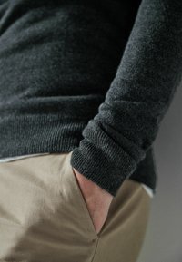 Ciemnoszary sweter dzianinowy z ściągaczem, noszony na białej koszuli i zestawiony z beżowymi spodniami. Widoczna ręka spoczywająca w kieszeni.