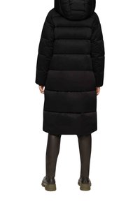 Schwarze Daunenjacke mit Kapuze, horizontalem Steppmuster, langen Ärmeln und knielanger Passform. Tragen mit schwarzen Strumpfhosen und klobigen Stiefeln.