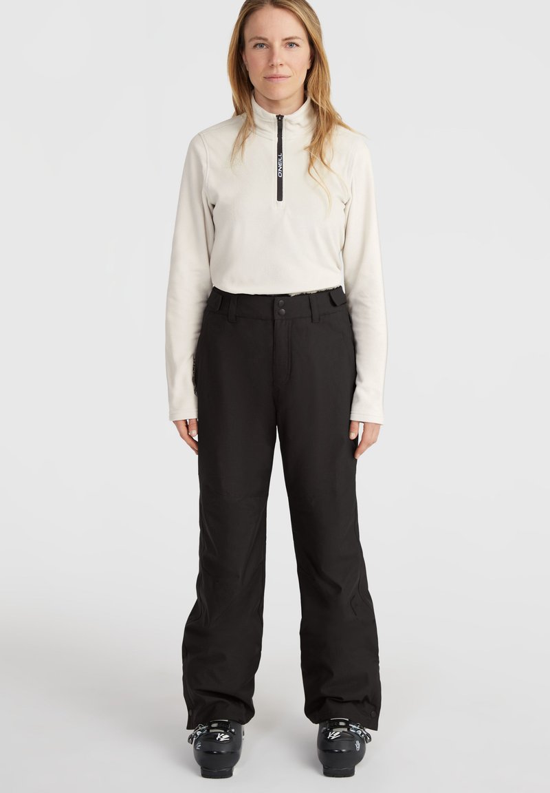 Leichtes, cremefarbenes Fleece-Oberteil mit Reißverschluss, kombiniert mit locker sitzenden, schwarzen Skihosen. Schwarze Skischuhe runden das Outfit ab.