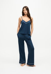 Femme debout pieds nus portant un haut sans manches bleu marine et un pantalon large assorti avec un liseré blanc sur un fond uni.