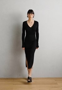 Robe noire à manches longues avec un décolleté en V profond, texture côtelée et fentes latérales, associée à des sandales à plateforme noires sur un sol en bois.