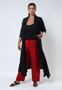 Lange zwarte cardigan over een zwarte tanktop en rode wijde broek, met een ontspannen pasvorm, zijzakken en platte zwarte sandalen.