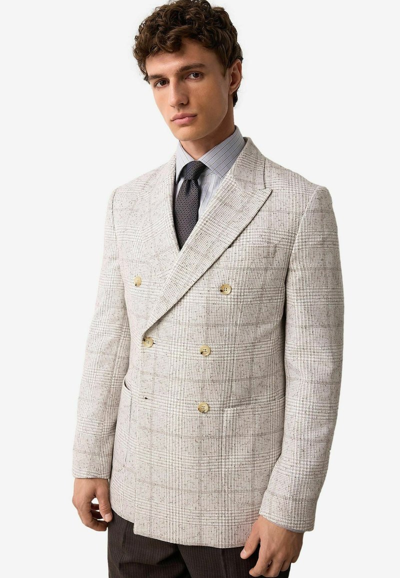 Kétgombos blézer világosszürke houndstooth mintával, arany gombokkal és textúrált anyagfelülettel. Kockás inggel párosítva.