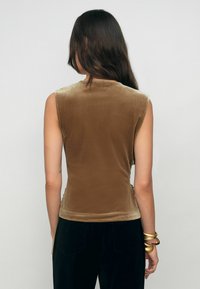 Barna bársonyos, ujjatlan felső, amely formázott kialakítású, sima tapintású és letisztult nyakkivágással rendelkezik. Magas derekú fekete nadrággal viselve.