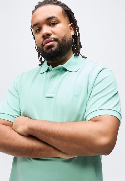 Άνδρας με κοτσίδες στα μαλλιά και μούσι φοράει ανοιχτό πράσινο πόλο μπλουζάκι, στέκεται με τα χέρια σταυρωμένα μπροστά σε μια μονόχρωμη ανοιχτή επιφάνεια.