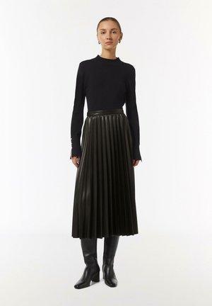 Schwarzes, geripptes Langarmshirt kombiniert mit einem hoch taillierten, plissierten Lederrock in Dunkelgrün und schwarzen kniehohen, Absatzstiefeln.