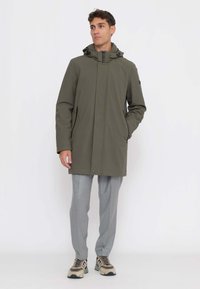 Cappotto impermeabile verde oliva con cappuccio, due tasche laterali con cerniera e chiusura frontale con zip. Il modello indossa pantaloni grigi e sneakers.