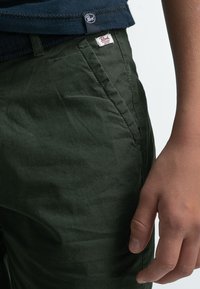 Pantaloni in tessuto leggero di colore verde scuro con una texture liscia. I dettagli notevoli includono un piccolo logo e una cucitura di tasca laterale.