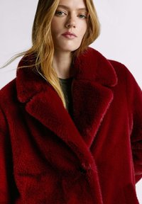 Cappotto in pelliccia sintetica rossa con colletto ampio, texture morbida e due bottoni. Il cappotto ha una vestibilità ampia ed è indossato sopra una camicia scura.