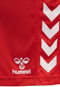 Shorts de sport rouges en tissu lisse, avec des motifs en chevrons blancs sur les côtés et un logo stylisé avec "hummel" en bas.