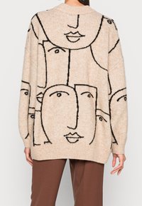 Beige oversized tröja med ett svart ansiktsmönster, ribbade ärmslut och nederkant. Mjuk textur, avslappnad passform och bred halsringning för en avslappnad stil.