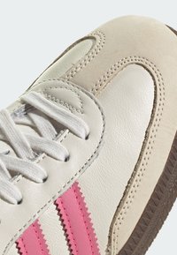 Sneaker in pelle bianca con strisce seghettate rosa, dotata di un accento in suede beige testurizzato, punta rotonda e lacci bianchi su uno sfondo chiaro.