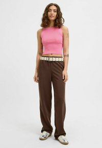 Rožinė raštuota trumpa palaidinė, rudos plačios kelnės su grietinėlės spalvos diržu, puoštu juodais taškeliais, ir sidabriniai sportbačiai. Neutralus fonas.