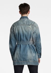Chaqueta de mezclilla en azul claro con desgastes, parches rasgados, cintura ceñida con cinturón y mangas largas. Presenta un cuello clásico y costura en el yugo posterior.