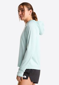 Vaaleansininen, pitkähihaisen hupullinen huppari kosteutta siirtävästä kankaasta, jossa on huppu ja peukaloaukot; yhdistetty mustiin shortseihin, joissa on logoyksityiskohta.