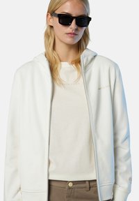 Weißer Reißverschluss-Hoodie mit glatter Textur, grauen Akzenten, kombiniert mit einem hellbeigen T-Shirt und braunen Hosen. Schwarze Sonnenbrille vorhanden.
