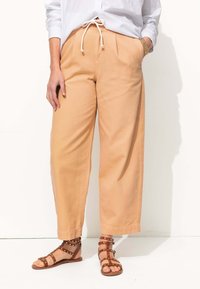 Pantalons larges couleur pêche clair avec des plis, taille élastique à cordon, et poches latérales, associés à des sandales marron à clous. Tissu en coton lisse.