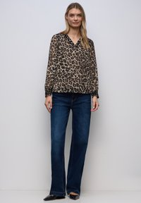 Ung kvinde iført leopardmønstret bluse med lange ærmer, mørkeblå jeans med lige ben og sorte spidse sko, stående mod en lysegrå baggrund.