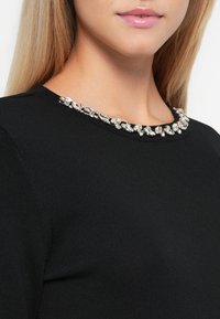 Detailný záber osoby v čiernom topy s okrúhlym výstrihom ozdobeným veľkými jasnými kryštálovými detailmi.
