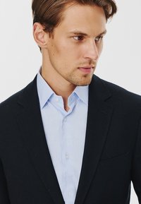 Granatowa marynarka nałożona na jasnoniebieską koszulę. Marynarka ma wyraźny kołnierz i kieszeń z przodu, a koszula charakteryzuje się klasycznym zapięciem na guziki.