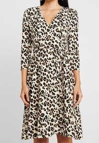 Sukienka typu wrap w lekkim materiale z kremowym tłem i czarnymi oraz brązowymi plamkami w leopardzie, z długimi rękawami i wiązanym detalem w talii.