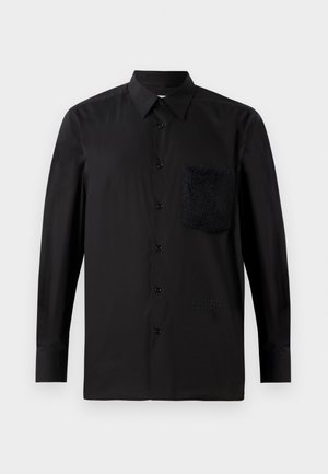 Chemise noire à manches longues avec une poche texturée foncée, fermée par des boutons, et un détail de marque subtil près de l'ourlet. Tissu lisse.