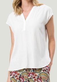 Haut blanc à manches courtes avec un décolleté en V, fabriqué en tissu léger. S'associe avec un pantalon à motifs présentant des designs floraux colorés.