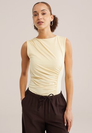 Žena s kučeravými vlasmi, nosiaca kruhové náušnice, žltý vrchný diel bez rukávov s riasením a tmavé nohavice so sťahovaním na šnúrku na neutrálnom pozadí.