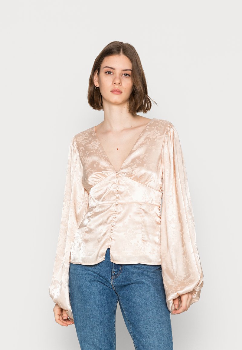 Blusa de cetim em pêssego claro, com mangas longas bufantes, decote em V profundo, fecho frontal com botões e padrão floral subtil. Combinada com jeans azuis.