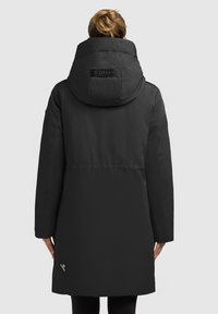 Svart puffjacka med en stor huva, med en texturerad yta, elastiska ärmslut och en dragkedjeficka nära fållen. Diskret logotyp på baksidan.