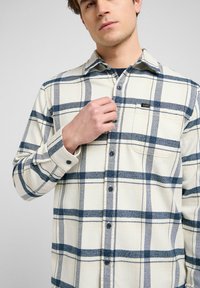 Camisa de cuadros de manga larga en color crema con rayas azules y marinas, cierre de botones en la parte delantera, un bolsillo en el pecho y diseño de cuello estándar.