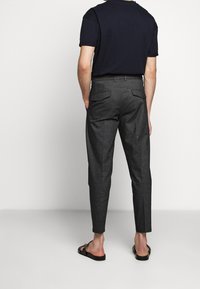 Man med mörkgrå skräddarsydda byxor med bakfickor, svarta sandaler och en svart kortärmad t-shirt, står mot en enfärgad bakgrund.