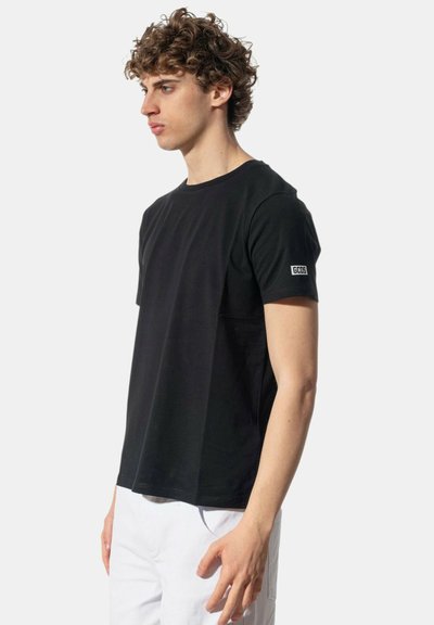 Giovane uomo con capelli ricci che indossa una t-shirt nera a maniche corte con un piccolo logo GCDS sulla manica e pantaloni bianchi, in piedi di lato.