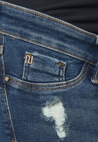 Vaqueros de mezclilla con un acabado desgastado, que presentan un desgarro en el bolsillo delantero. Se pueden ver detalles en cobre y costuras en contraste.