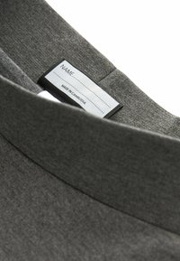 Grijze stoffen kraag van het kledingstuk met een label waarop staat "NAAM: GEMAAKT IN CAMBODJA." Het materiaal heeft een gestructureerde afwerking, met een gladde rand.