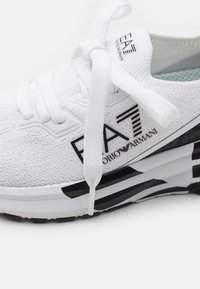 Valkoinen urheilukenkä, jossa on teksturoitu neulottu päällinen, mustat yksityiskohdat ja muotoiltu pohja, jossa on mustavalkoiset raidat.