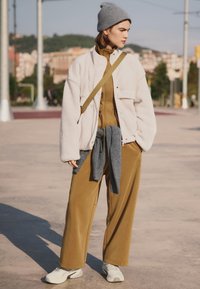 Übergroße beige Fleecejacke, olivgrüne Reißverschlussjacke zum Layering, graue Strickmütze, grauer Pullover um die Taille gebunden und cremefarbene Sneaker.