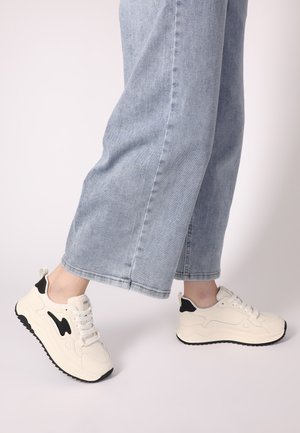 Weiße Sneaker mit schwarzen Akzenten, glatten Materialien, runder Form und strukturiertem Sohle, kombiniert mit hellblauen, weiten Jeans.