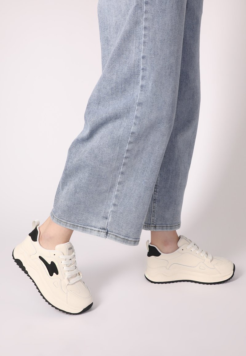 Weiße Sneaker mit schwarzen Akzenten, glatten Materialien, runder Form und strukturiertem Sohle, kombiniert mit hellblauen, weiten Jeans.