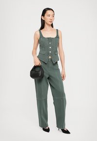 Zeleno denim brezrokavnik z gumbi in dekorativnimi žebljički, v kombinaciji s širokimi zelenimi hlačami. Model drži majhno črno torbico.