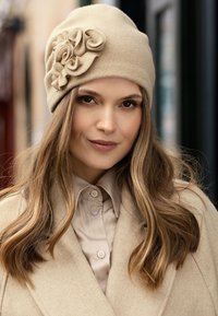 Béret en laine beige avec un accent floral en tissu, présentant une texture douce et un ajustement serré. Porté avec un manteau et une chemise beige clair.