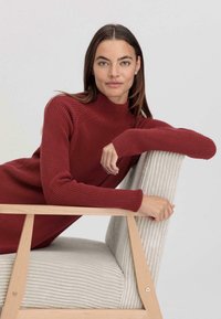 Maglione a collo alto a coste rosso con maniche lunghe, abbinato a una sedia in tessuto beige a coste con struttura in legno.