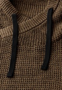Hnědý pletený svetr s texturovaným vzorem, který má černou stahovací šňůrku u límce. Tkanina vypadá silně a robustně.