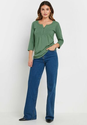 Groene lange mouw top met een V-hals, geplooide voorkant en driekwartmouwen, gecombineerd met blauwe wijde jeans en zwarte puntige hakken.