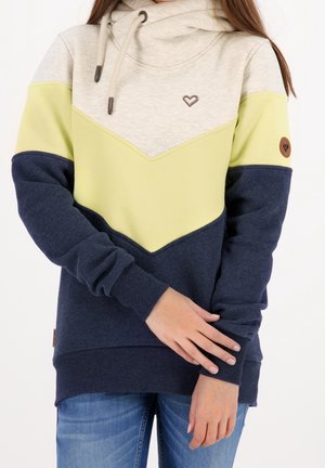 Femme portant un hoodie color-block beige, jaune et bleu marine, avec un petit logo en forme de cœur sur la poitrine et des manches retroussées.