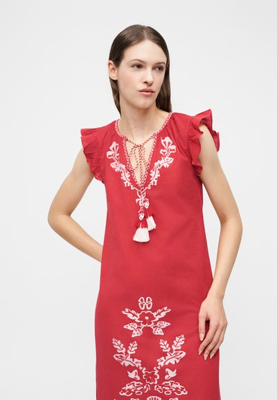 Γυναίκα που φοράει αμάνικο κόκκινο φόρεμα με λευκή κεντητή floral διακόσμηση, μανίκια καπέλου με βολάν και κορδόνια με φουντίτσες στον λαιμό, στέκεται μπροστά σε λευκό φόντο.